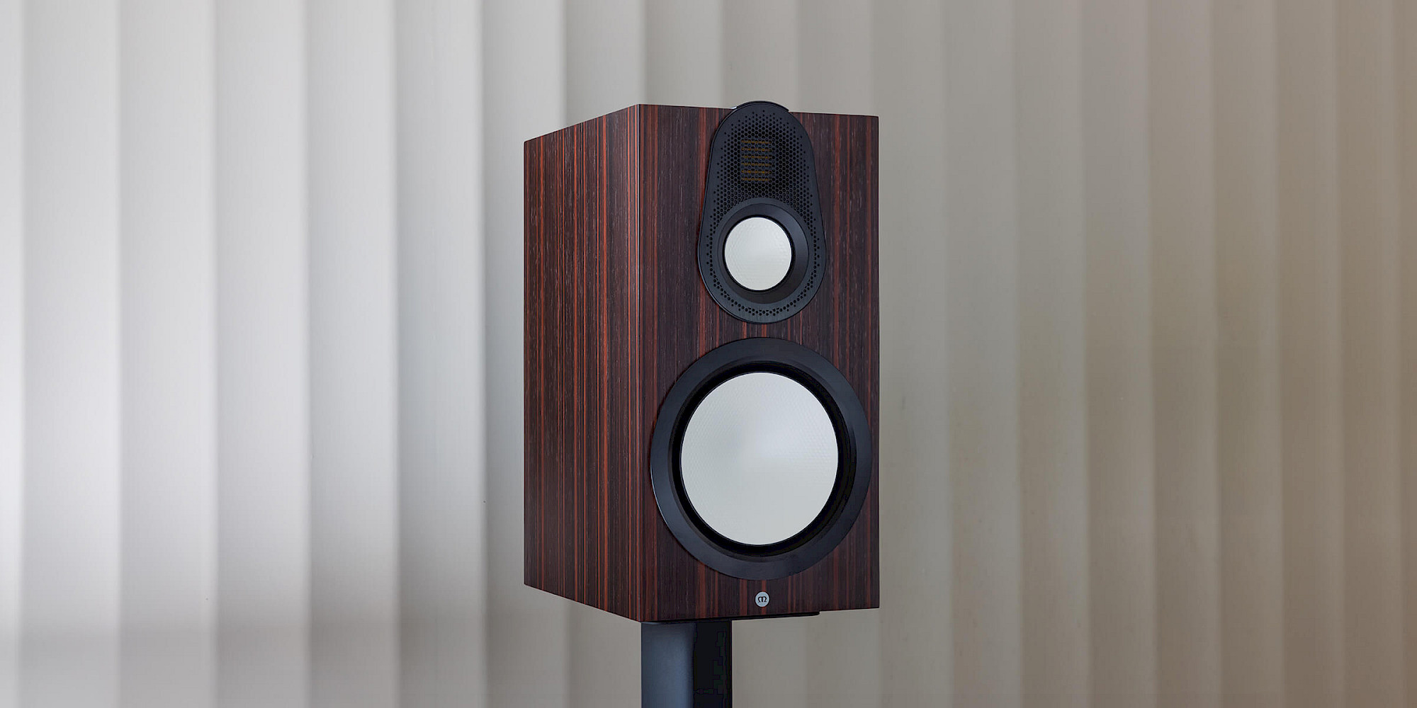 Closeup of the Monitor Audio Gold 100 6G bookshelf speaker in Macassar Wood Veneer finish, placed on a black stand.
